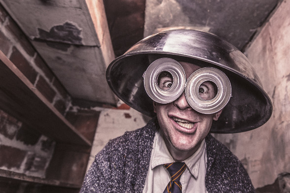 A man in an abandoned building wearing a suit and tie, a colander, and silly glasses, making a silly face.