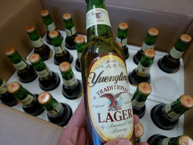 Someone holding an unopened bottle of Yuengling lager, with an entire case in the background.