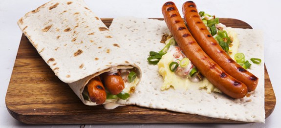 Two tunnbrödsrullar sitting next to each other on a wooden cutting board, one wrapped up and one open to display the contents (two grilled hotdogs, mayo, and salad).