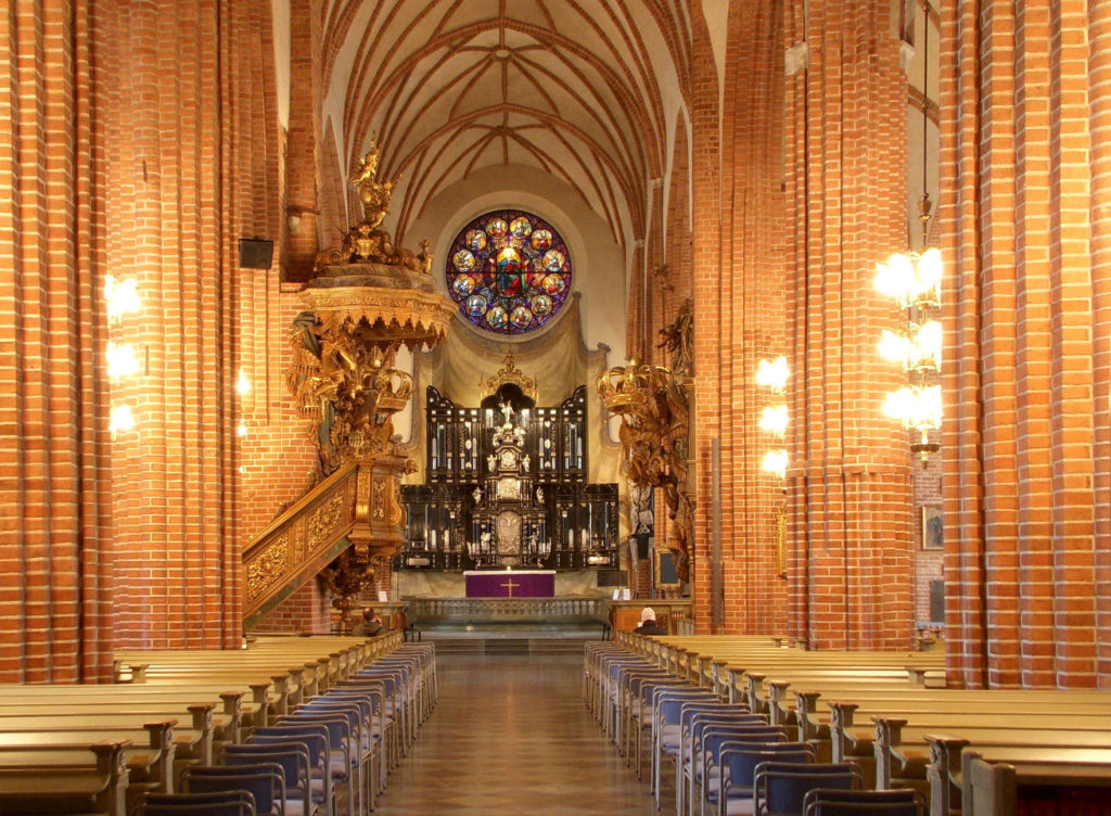 The interior of Storkyrkan in Stockholm, Sweden. The view is down the center aisle, facing a stained glass rossette. On the left hand side is a spiral staircase attached to a column, leading to a pulpit. The ceilings are high and vaulted; the columns are red brick. The seats on either side are empty.