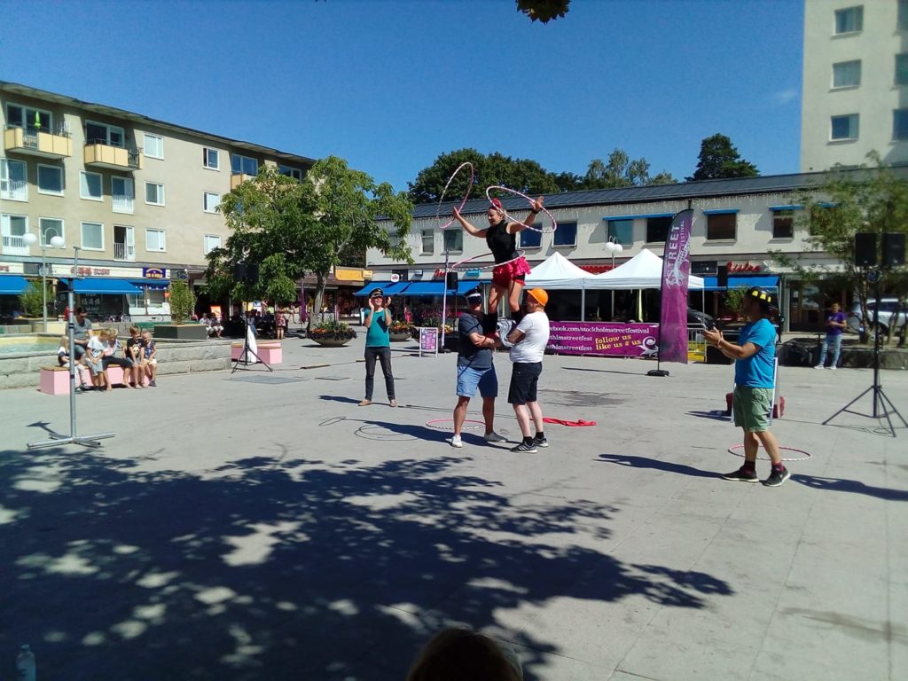 A hula hooping street performer standing on the shoulders of two men, spinning one hoop around her waist and one around each arm.