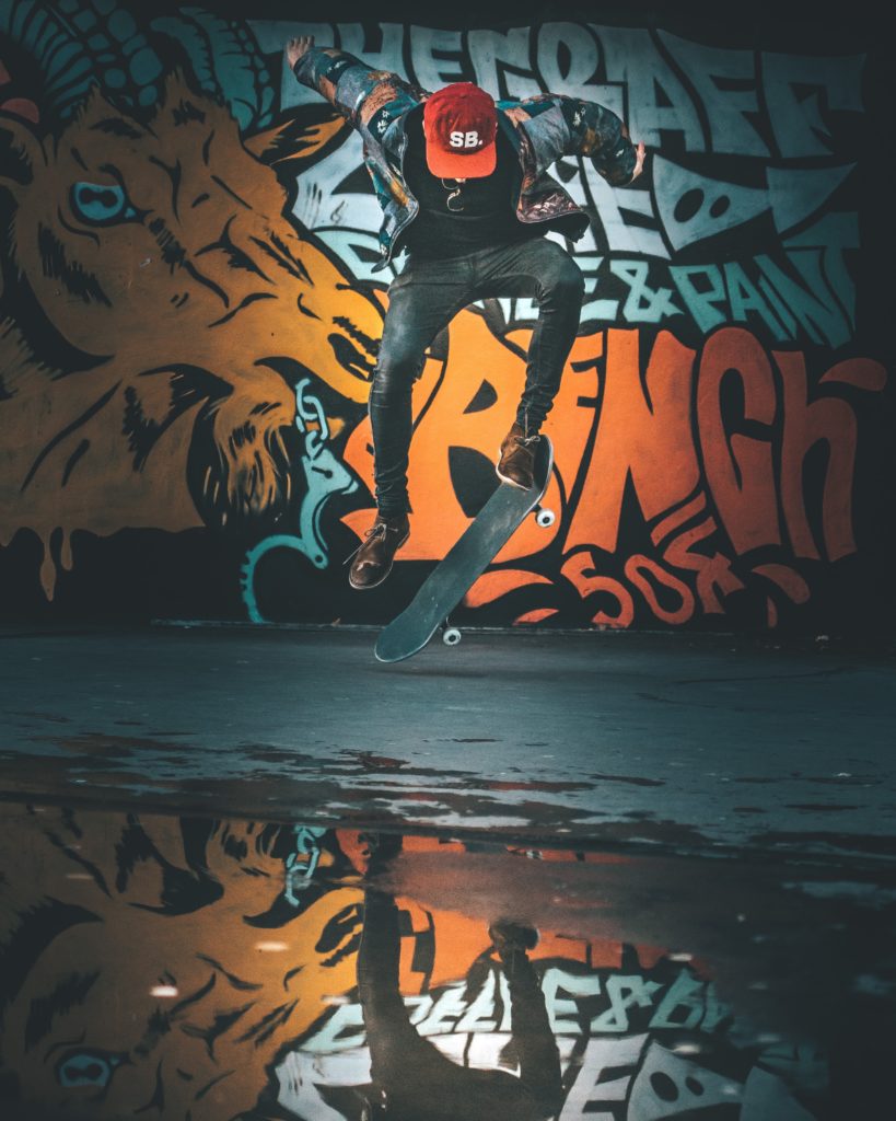 A man performing a skateboard trick in front of orange graffiti on a wall. His face is obscured by a red baseball cap and he's partially reflected in the puddle under him.
