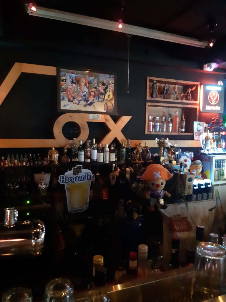 A dark bar, with "Cox" in light wood mounted on the black wall above the bottles.