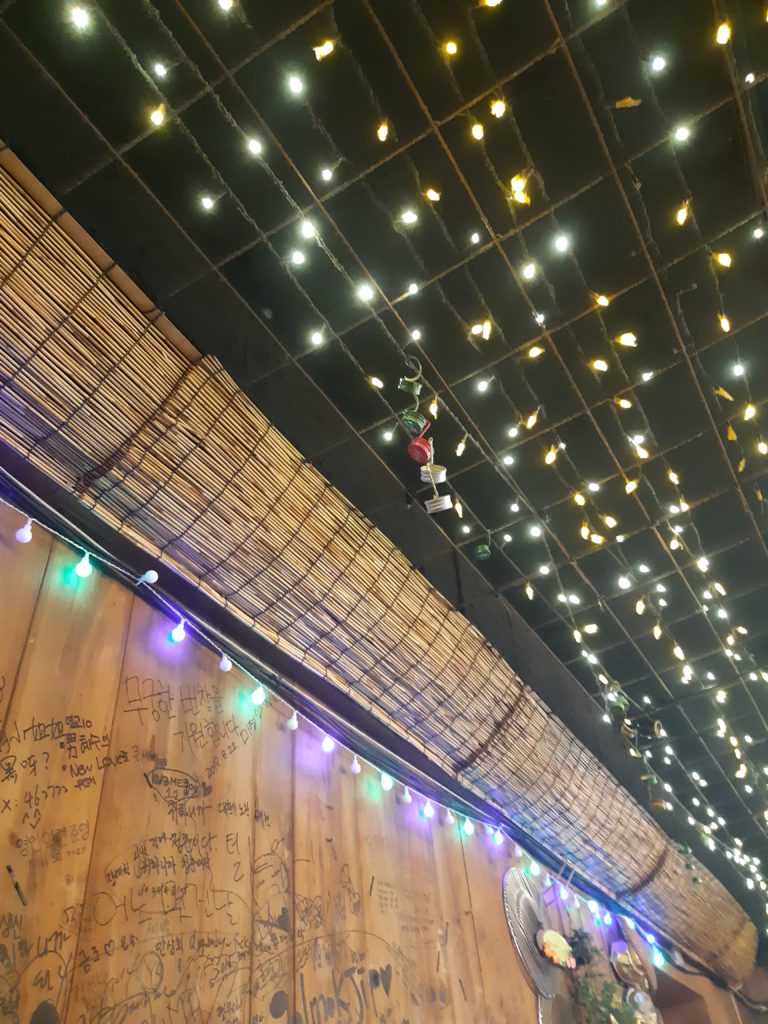 A chain of soju bottle caps dangling from a ceiling decorated in fairy lights.