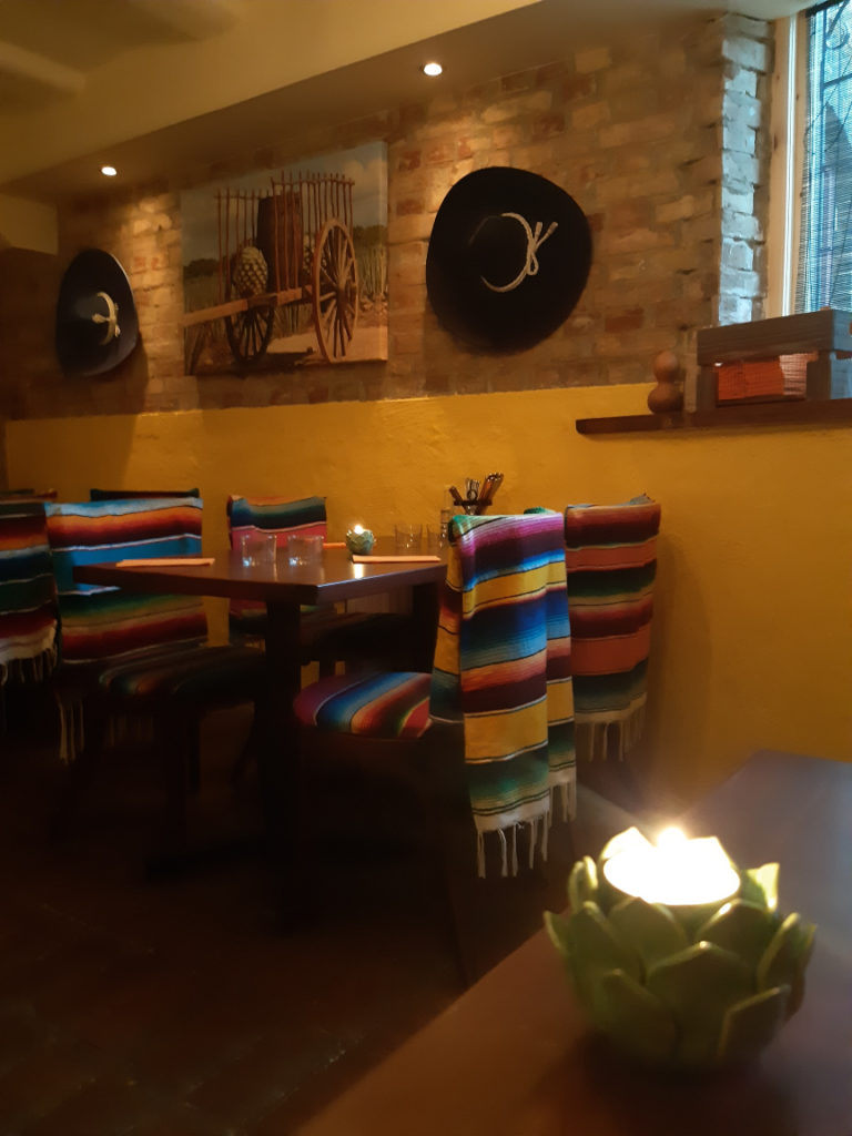 Interior shot of an empty Mexican restaurant with bright yellow walls, black sombreros hanging from the wall, and chairs upholstered in bright stripes.